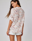 A woman with long, wavy brown hair stands facing away, smiling over her shoulder. She wears a light Bella Dahl Leinenmix Bluse mit Blumenprint and white shorts with rolled sleeves, set against a plain light gray background.