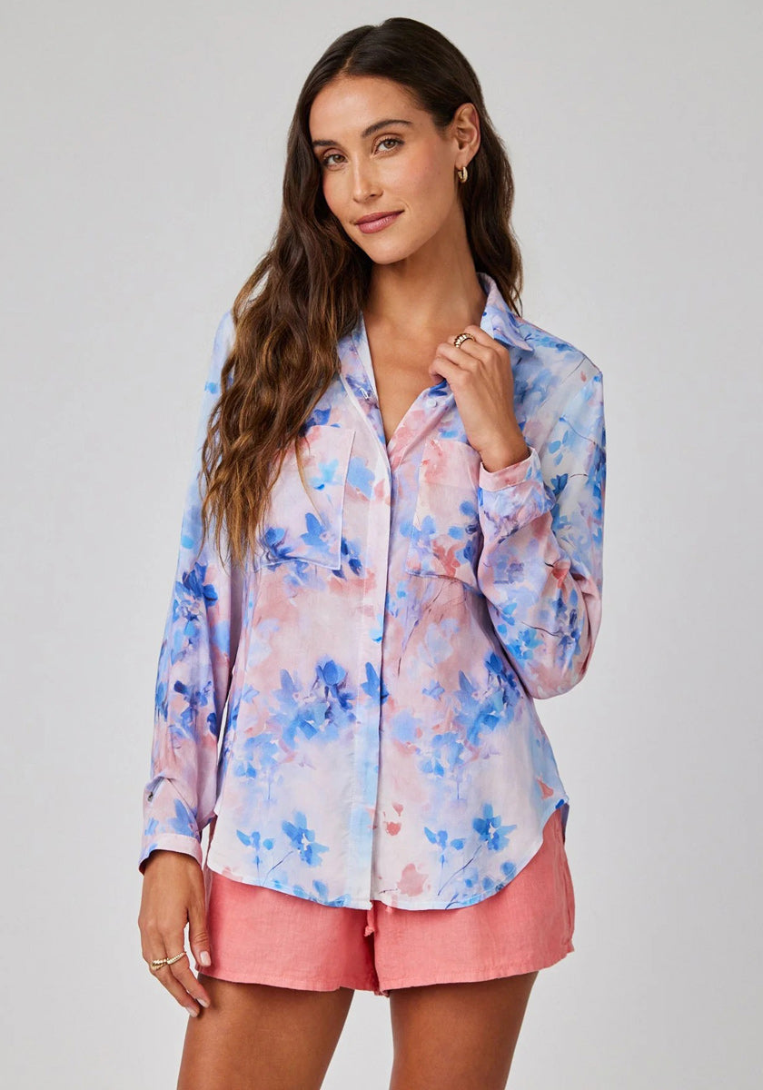 Eine Frau mit langen, welligen braunen Haaren trägt eine lockere Floral-Print Hemdbluse mit langen Ärmeln in Pink und Blau von Bella Dahl, kombiniert mit korallrosanen Shorts. Sie steht vor einem hellgrauen Hintergrund und lächelt sanft.