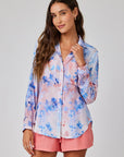 Eine Frau mit langen, welligen braunen Haaren trägt eine lockere Floral-Print Hemdbluse mit langen Ärmeln in Pink und Blau von Bella Dahl, kombiniert mit korallrosanen Shorts. Sie steht vor einem hellgrauen Hintergrund und lächelt sanft.
