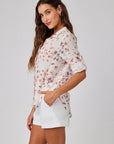 A woman in profile with long wavy brown hair and a slight smile wears a Bella Dahl Leinenmix Bluse with pink and orange Blumenprint and white shorts, left hand in her pocket, standing against a plain background.
