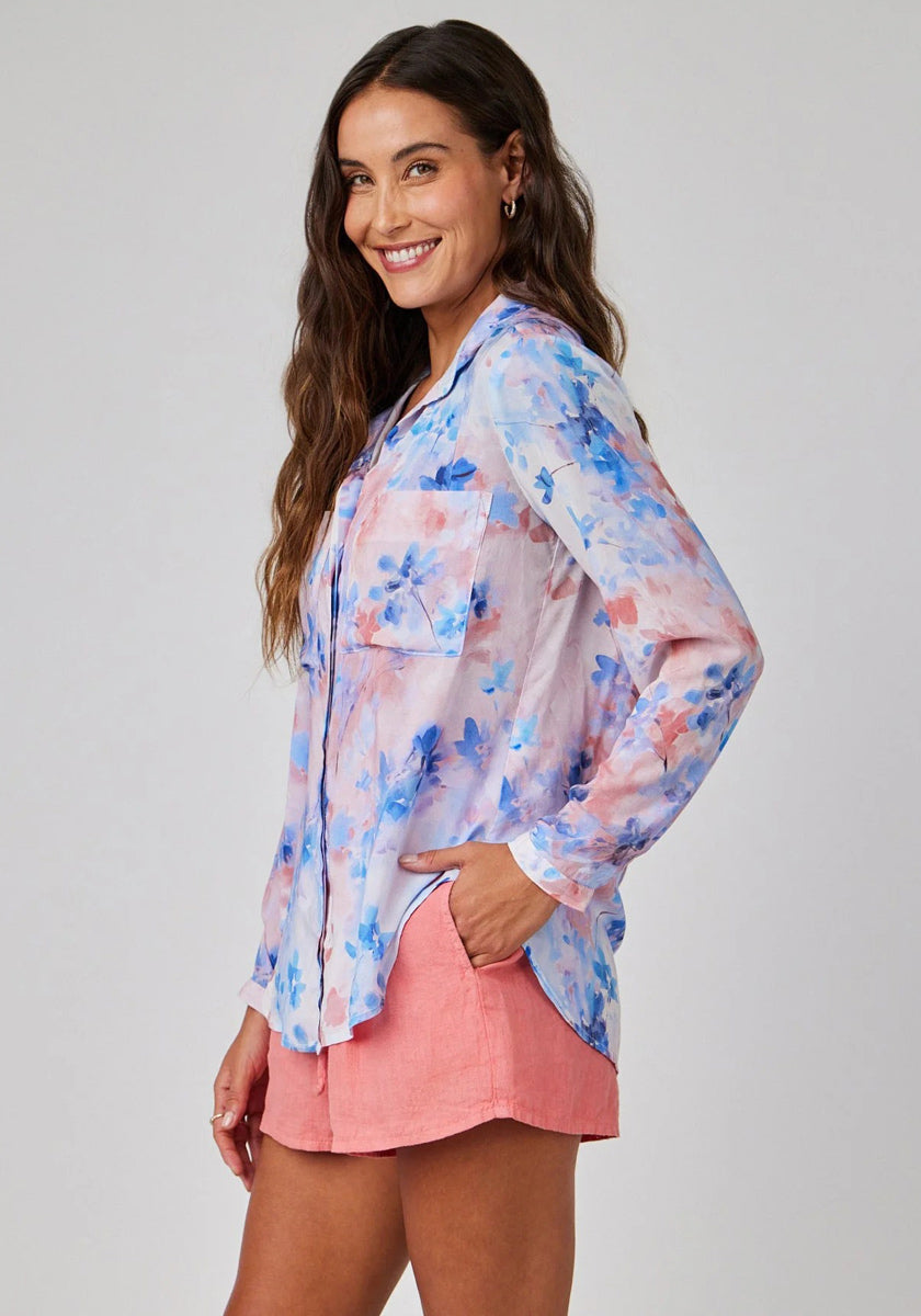 A woman with long, wavy brown hair smiles while wearing a sheer Bella Dahl Hemdbluse mit langen Ärmeln in blue and pink floral print, paired with coral pink shorts. She stands sideways with a hand in her pocket against a light gray background.
