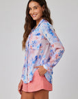 A woman with long, wavy brown hair smiles while wearing a sheer Bella Dahl Hemdbluse mit langen Ärmeln in blue and pink floral print, paired with coral pink shorts. She stands sideways with a hand in her pocket against a light gray background.