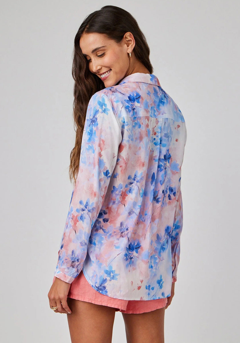 A woman with long brown hair and gold hoop earrings smiles over her shoulder, wearing a Bella Dahl floral-print Hemdbluse with long sleeves in blue, pink, and white tones, paired with pink shorts. The background is plain light gray.