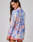 A woman with long brown hair and gold hoop earrings smiles over her shoulder, wearing a Bella Dahl floral-print Hemdbluse with long sleeves in blue, pink, and white tones, paired with pink shorts. The background is plain light gray.