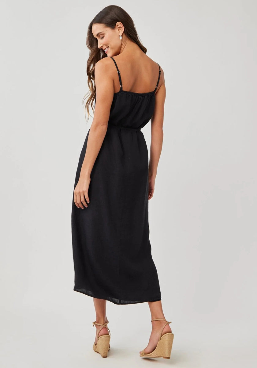 A woman with long brown hair wears a sleeveless black Bella Dahl linen midi dress with adjustable straps and tan wedge sandals, slightly turned and smiling over her shoulder against a plain light background.