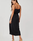 A woman with long brown hair wears a sleeveless black Bella Dahl linen midi dress with adjustable straps and tan wedge sandals, slightly turned and smiling over her shoulder against a plain light background.