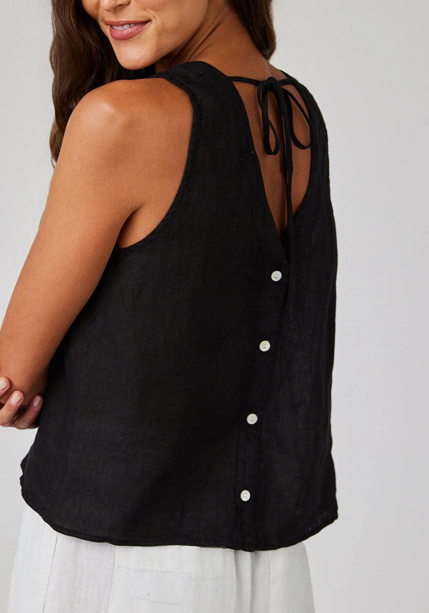 A woman wears a sleeveless Bella Dahl linen top with tie straps and white buttons down the back, paired with light-colored pants. She smiles, turned slightly to the side, her long brown hair resting over one shoulder.