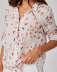 A woman models a light, semi-sheer Bella Dahl Leinenmix Bluse featuring a loose fit and delicate pink, orange, and gray Blumenprint. She holds the hem of the Hemdbluse with one hand, standing against a plain background.