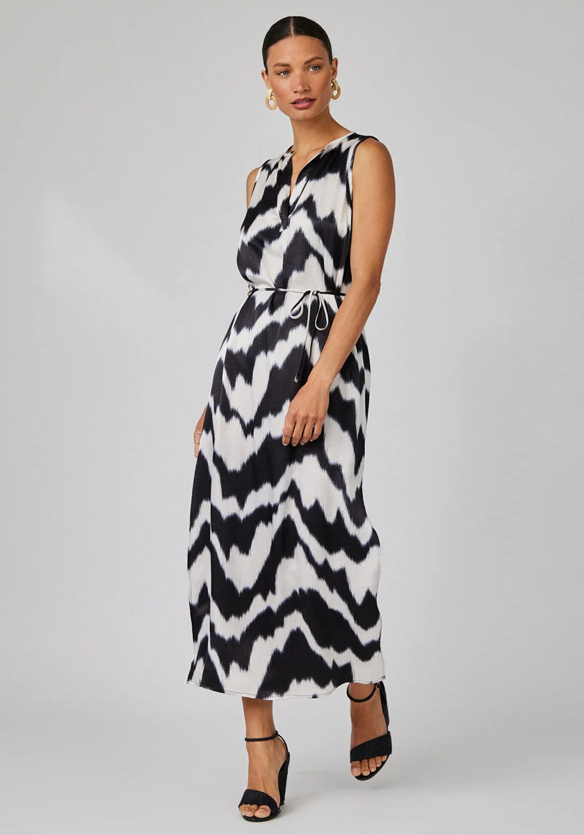A woman in a Bella Dahl sleeveless midi dress with abstract print and tie belt stands against a plain background. She has her hair pulled back, wears large gold hoop earrings and black heeled sandals, her expression neutral.