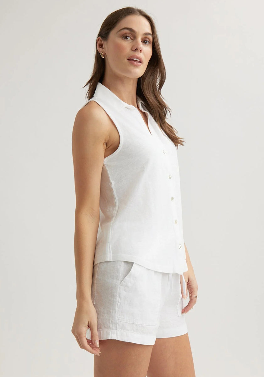 A woman with long brown hair models a sleeveless white button-up shirt with Hemdkragen and white shorts from Bella Dahl, standing against a plain light background. She faces slightly to the side, looking confidently at the camera.