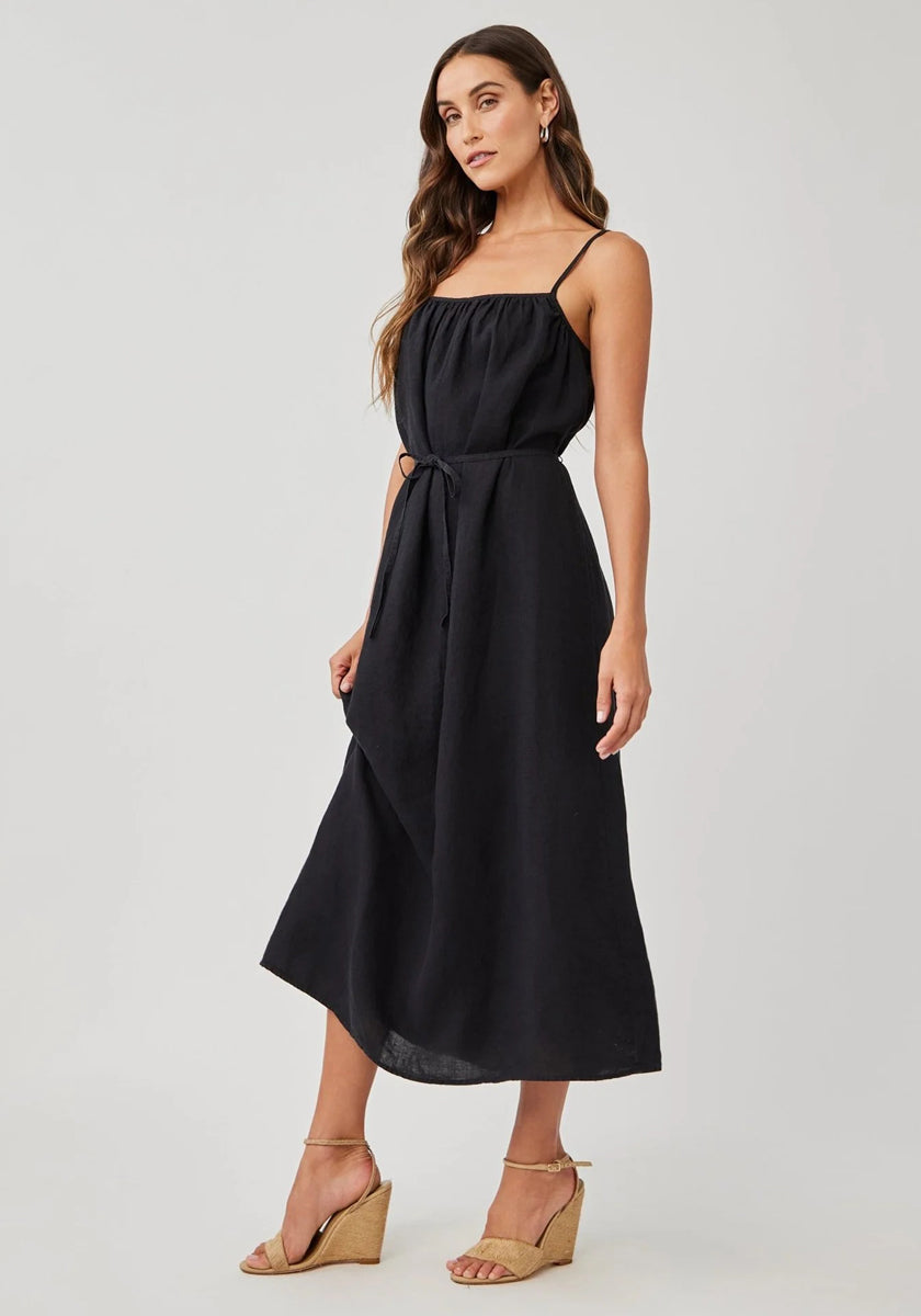 A woman poses sideways in a studio wearing a Bella Dahl Leinen Midikleid with verstellbaren Trägern and Bindegürtel, styled with gold hoop earrings and beige Keilsandalen. She has long brown hair and stands against a plain background.
