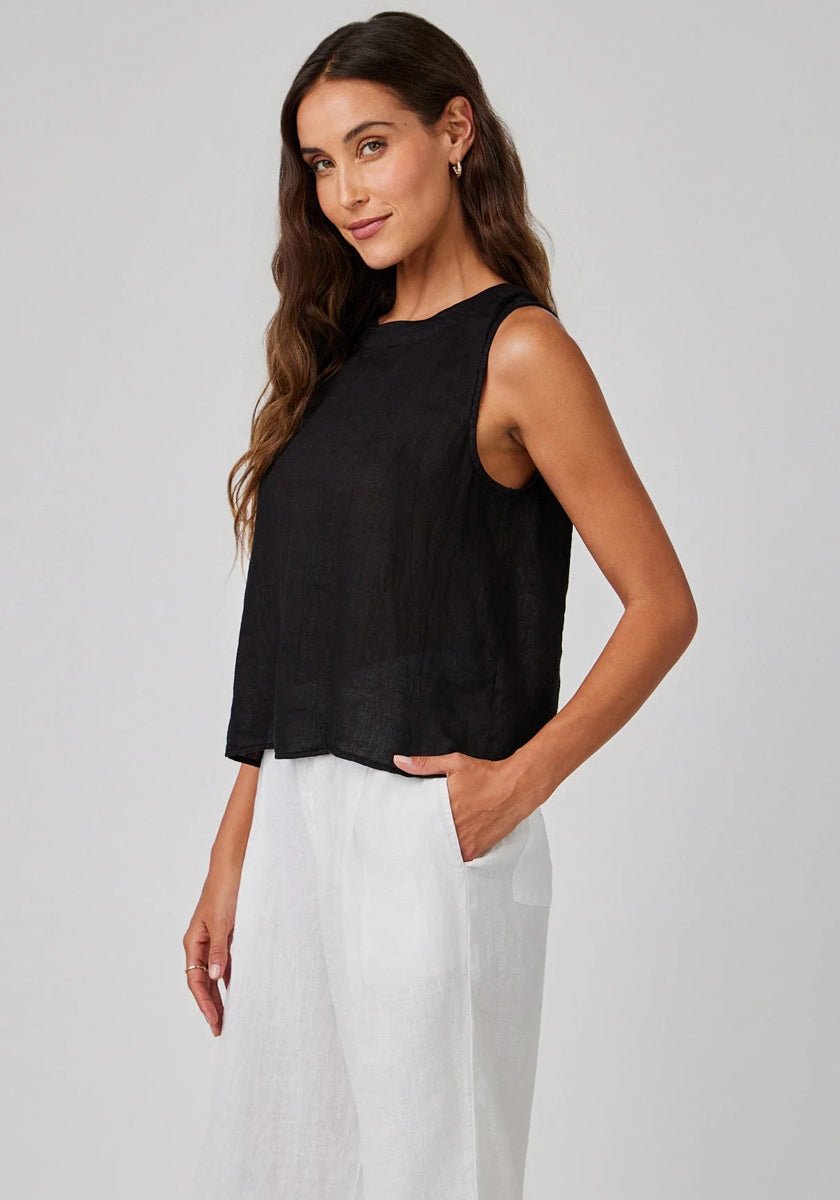 A woman with long wavy brown hair wears a sleeveless Bella Dahl linen top and loose white pants, standing sideways with one hand in her pocket, smiling slightly against a plain light gray background.