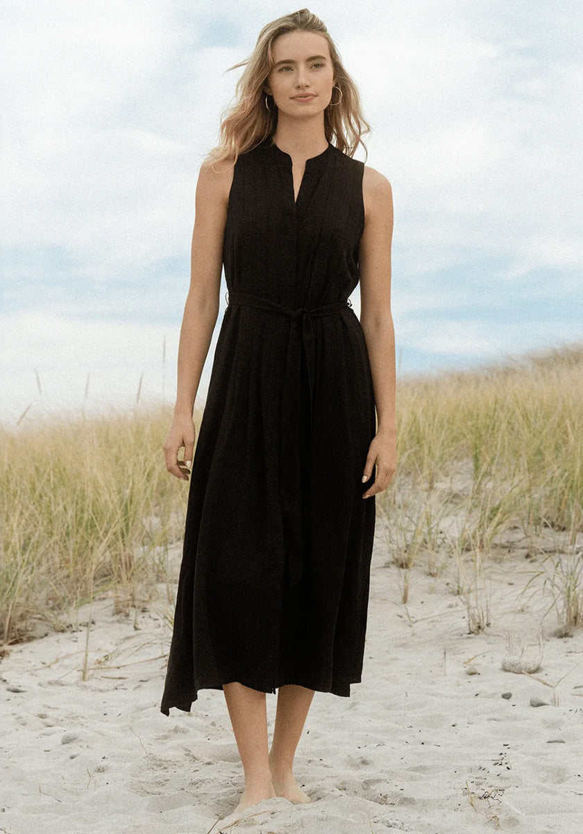 A woman with light skin and long wavy hair stands barefoot on sandy ground with tall grass, wearing a Bella Dahl black Lyocell midi dress with a tie belt. She gazes softly ahead beneath a cloudy sky, creating a serene, natural scene.
