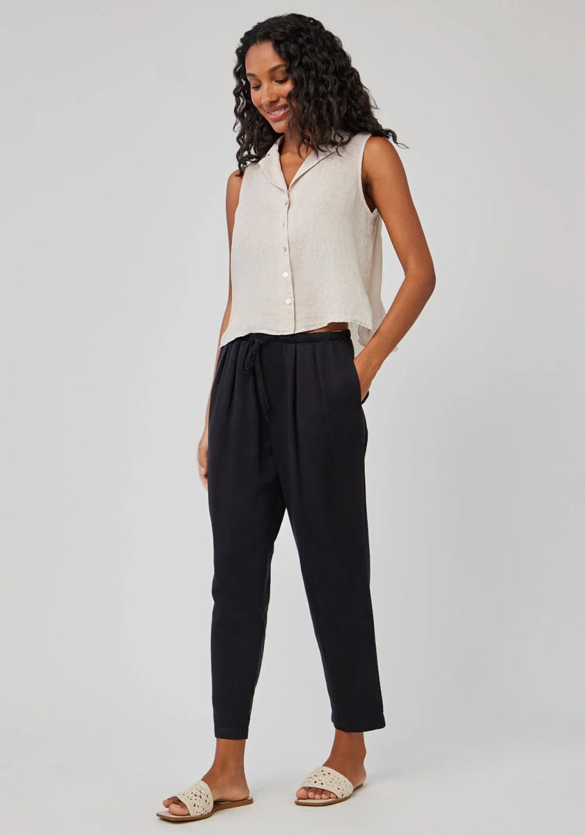 A woman with long curly hair wears a sleeveless beige button-up top and Bella Dahl black Lyocell-Leinen 7/8-length pants with an elastic waistband, paired with light slide sandals. She stands smiling, one hand in her pocket, against a light gray background.