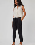 A woman with long curly hair wears a sleeveless beige button-up top and Bella Dahl black Lyocell-Leinen 7/8-length pants with an elastic waistband, paired with light slide sandals. She stands smiling, one hand in her pocket, against a light gray background.