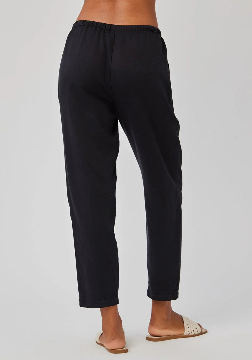 A person stands facing away, wearing Bella Dahls 7/8-length Lyocell-linen pants with an elastic waistband and light beige slide sandals. The plain, light-colored background shows only their lower back and legs.