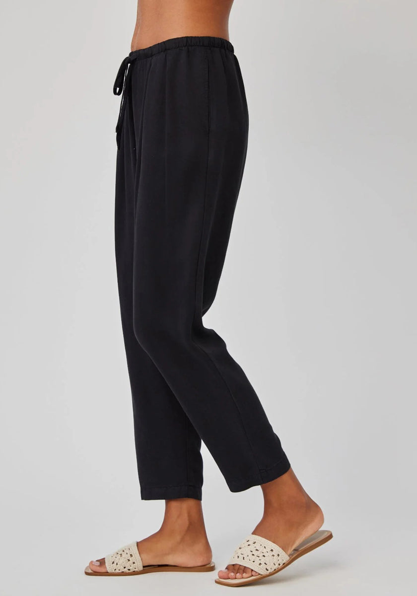 A person stands sideways against a light gray background, wearing Bella Dahl black Lyocell-Leinen-Hose in 7/8 length with elastic waistband and cream woven slide sandals. Only the lower body is visible, highlighting the casual, comfortable style.