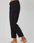 A person stands sideways against a light gray background, wearing Bella Dahl black Lyocell-Leinen-Hose in 7/8 length with elastic waistband and cream woven slide sandals. Only the lower body is visible, highlighting the casual, comfortable style.