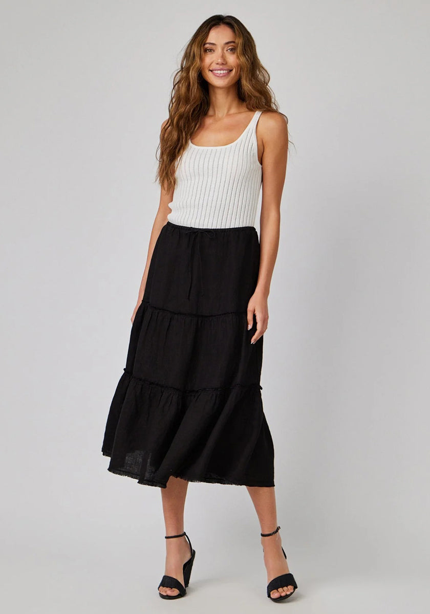 A woman with long wavy hair wears a white ribbed Bella Dahl tank top tucked into a black tiered midi skirt. She stands smiling, hands at her sides, and pairs the look with black open-toe heels against a light gray background.
