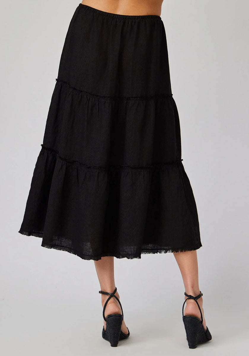 A woman is shown from the waist down wearing a black, tiered Bella Dahl Leinen Midirock with a frayed hem and ankle length, paired with black strappy wedge sandals. The background is plain light gray.