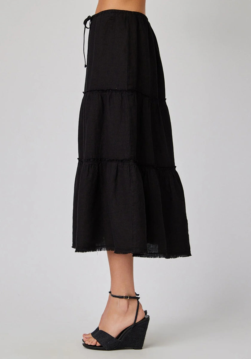 A person wears a black Leinen Midirock by Bella Dahl with Stufenschnitt, a drawstring waist, and raw hem, paired with black wedge sandals. The photo captures a side view against a plain light gray background.