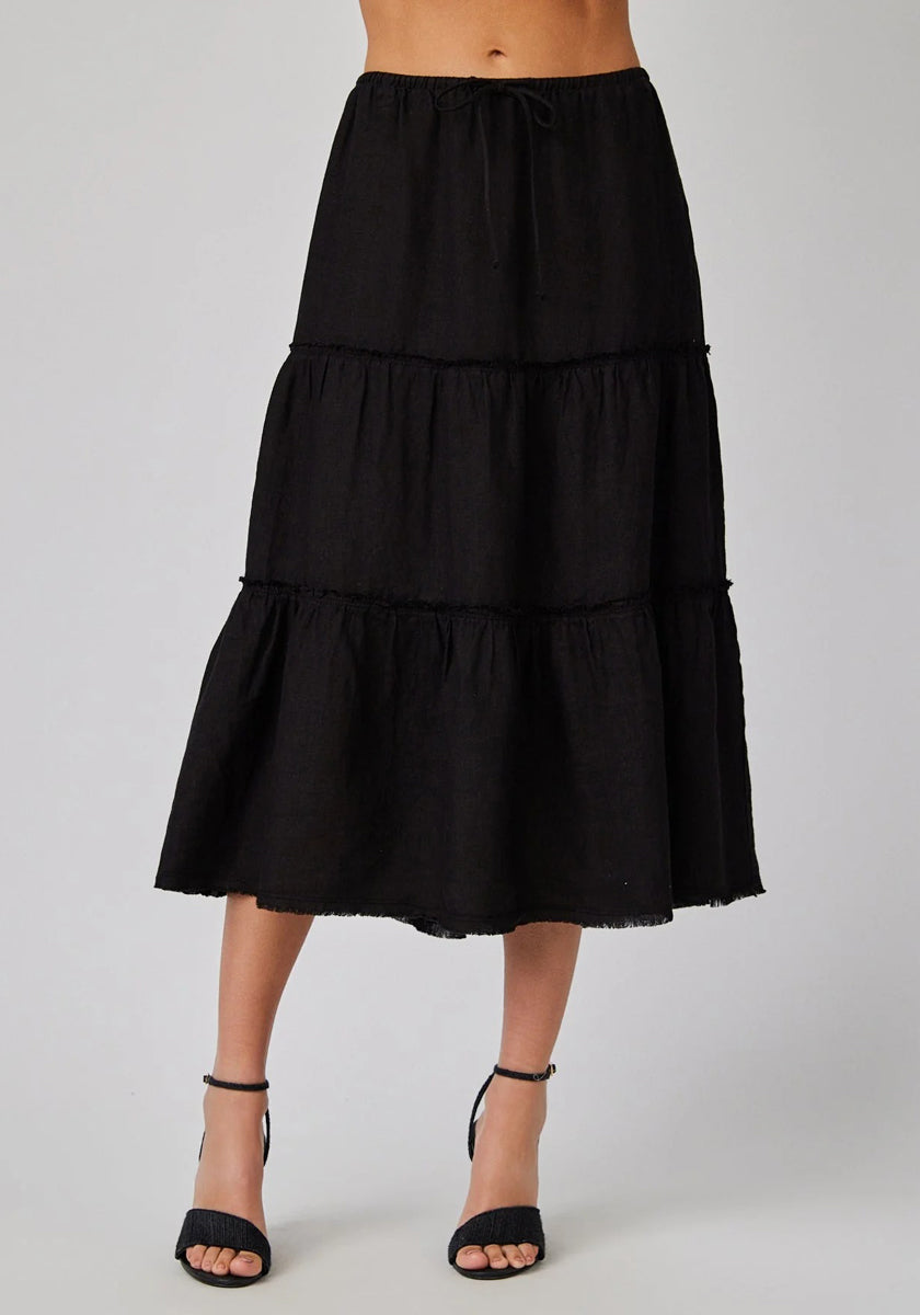 A person stands against a plain background, wearing a high-waisted black Bella Dahl Leinen Midirock with a Stufenschnitt featuring three ruffled tiers and a drawstring waistband, paired with black ankle-strap heeled sandals. The torso is not visible.