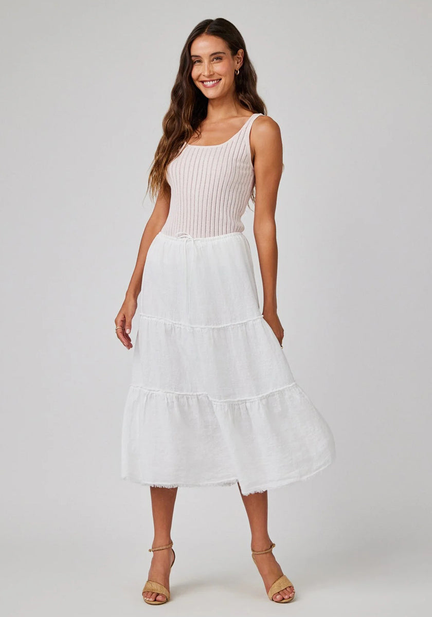A woman with long brown hair wears a light pink sleeveless ribbed top and a tiered white linen midi skirt by Bella Dahl. She smiles, posing in beige strappy heels against a plain light gray background.