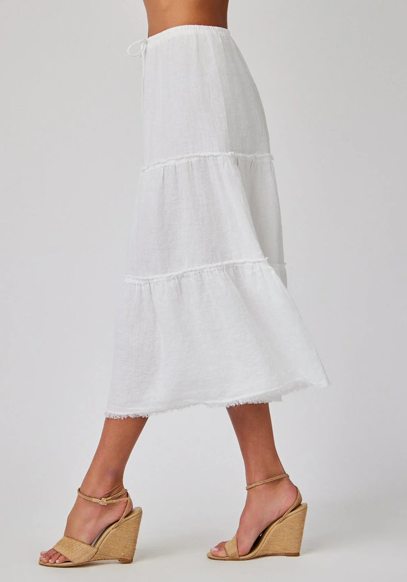 A person wears a high-waisted, white Bella Dahl midirock with stufenschnitt, crafted from light, semi-sheer linen and finished with a raw hem. They pair it with tan ankle-strap wedge sandals featuring braided jute soles against a plain background.