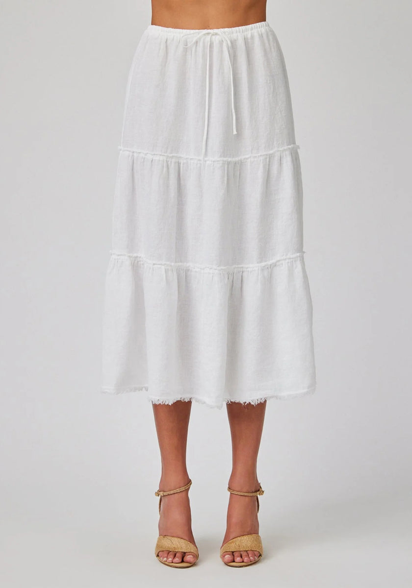 A person wears a white, tiered Bella Dahl linen midi skirt featuring a drawstring waist and frayed hem. The soft, textured fabric flows gracefully. Only the lower half is visible, styled with tan open-toe ankle-strap heels.