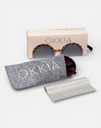 Eine schildpattfarbene runde Sonnenbrille von Okkia mit dunklen UV-400-Schutz-Gläsern ruht teilweise in einem grauen Filzetui, das passende Putztuch davor und ein offenes rosa Etui dahinter, das die Sonnenbrille präsentiert.