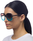 A woman with straight black hair in a low ponytail is shown in profile wearing Okkia gradient sunglasses—brown at the top and turquoise-blue at the bottom. She wears a white shirt against a plain white background.