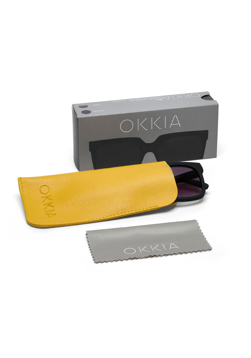 A pair of black square Okkia sunglasses is arranged on a white background with a yellow case, gray cleaning cloth, and gray Okkia-branded box. Both the case and cloth display the Okkia logo.