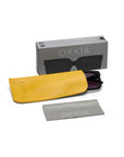 A pair of black square Okkia sunglasses is arranged on a white background with a yellow case, gray cleaning cloth, and gray Okkia-branded box. Both the case and cloth display the Okkia logo.