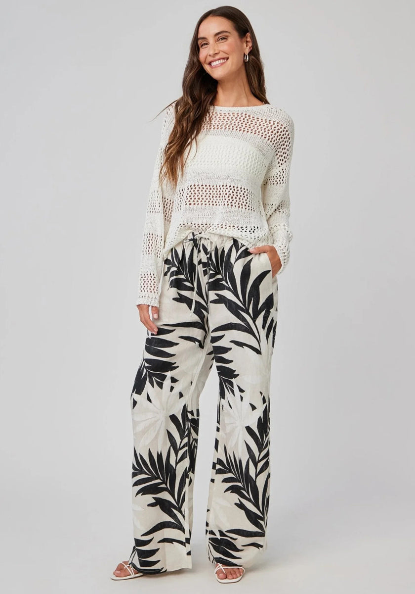 A woman smiles in a long-sleeve white crochet sweater and cream Bella Dahl linen pants with wide legs and palm print. With long wavy hair, hands in pockets, and sandals, she poses against a plain light gray background.