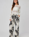 A woman smiles in a long-sleeve white crochet sweater and cream Bella Dahl linen pants with wide legs and palm print. With long wavy hair, hands in pockets, and sandals, she poses against a plain light gray background.