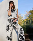A woman stands outdoors before a white wall, smiling warmly in a sleeveless white Bella Dahl jumpsuit with wide legs and bold black palm print; her wavy brown hair and green plants in the background add a fresh vibe.