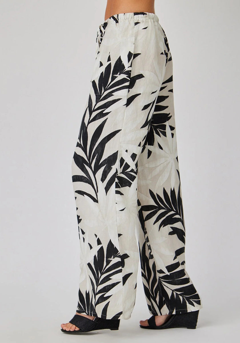 A person in profile wears a loose, high-waisted Bella Dahl Leinenhose with bold black Palmen-Print. The flowy, wide-leg pants are paired with open-toed black wedge sandals against a plain light background.