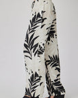 A person in profile wears a loose, high-waisted Bella Dahl Leinenhose with bold black Palmen-Print. The flowy, wide-leg pants are paired with open-toed black wedge sandals against a plain light background.