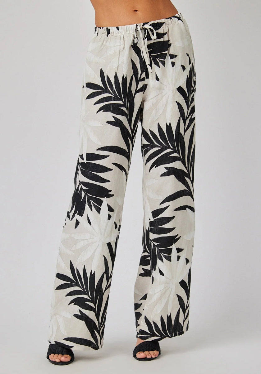 A person stands against a plain background, wearing Bella Dahl linen pants with a palm print and wide legs. The pants feature an elastic waistband with drawstring, paired with black open sandals. Upper body not visible.