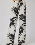 A person stands against a plain background, wearing Bella Dahl linen pants with a palm print and wide legs. The pants feature an elastic waistband with drawstring, paired with black open sandals. Upper body not visible.