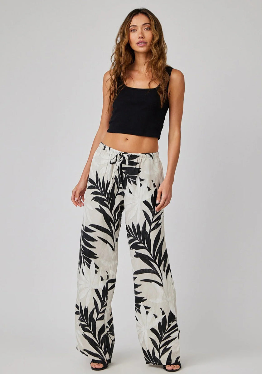A woman with long, wavy brown hair poses confidently against a light gray background, wearing a black sleeveless crop top and high-waisted Bella Dahl Leinenhose mit weitem Bein und Palmen-Print.