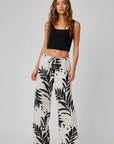 A woman with long, wavy brown hair poses confidently against a light gray background, wearing a black sleeveless crop top and high-waisted Bella Dahl Leinenhose mit weitem Bein und Palmen-Print.