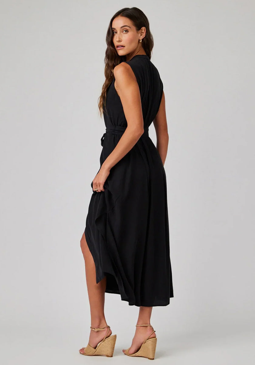 Eine Frau mit langem braunen Haar trägt ein schwarzes Midikleid von Bella Dahl aus Lyocell mit Bindegürtel, steht seitlich und blickt über die Schulter. Beige Keilsandaletten ergänzen ihren Look vor hellgrauem Hintergrund.