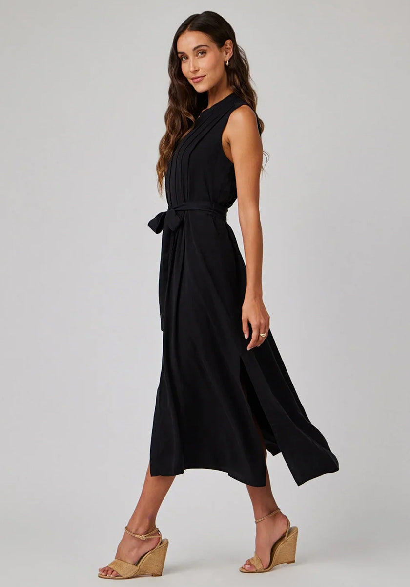 A woman poses sideways in a sleeveless, black Bella Dahl Midikleid made from soft Lyocell with a high neckline, waist tie, and side slit. She wears tan wedge sandals, has long wavy brown hair, and minimal jewelry against a light grey background.