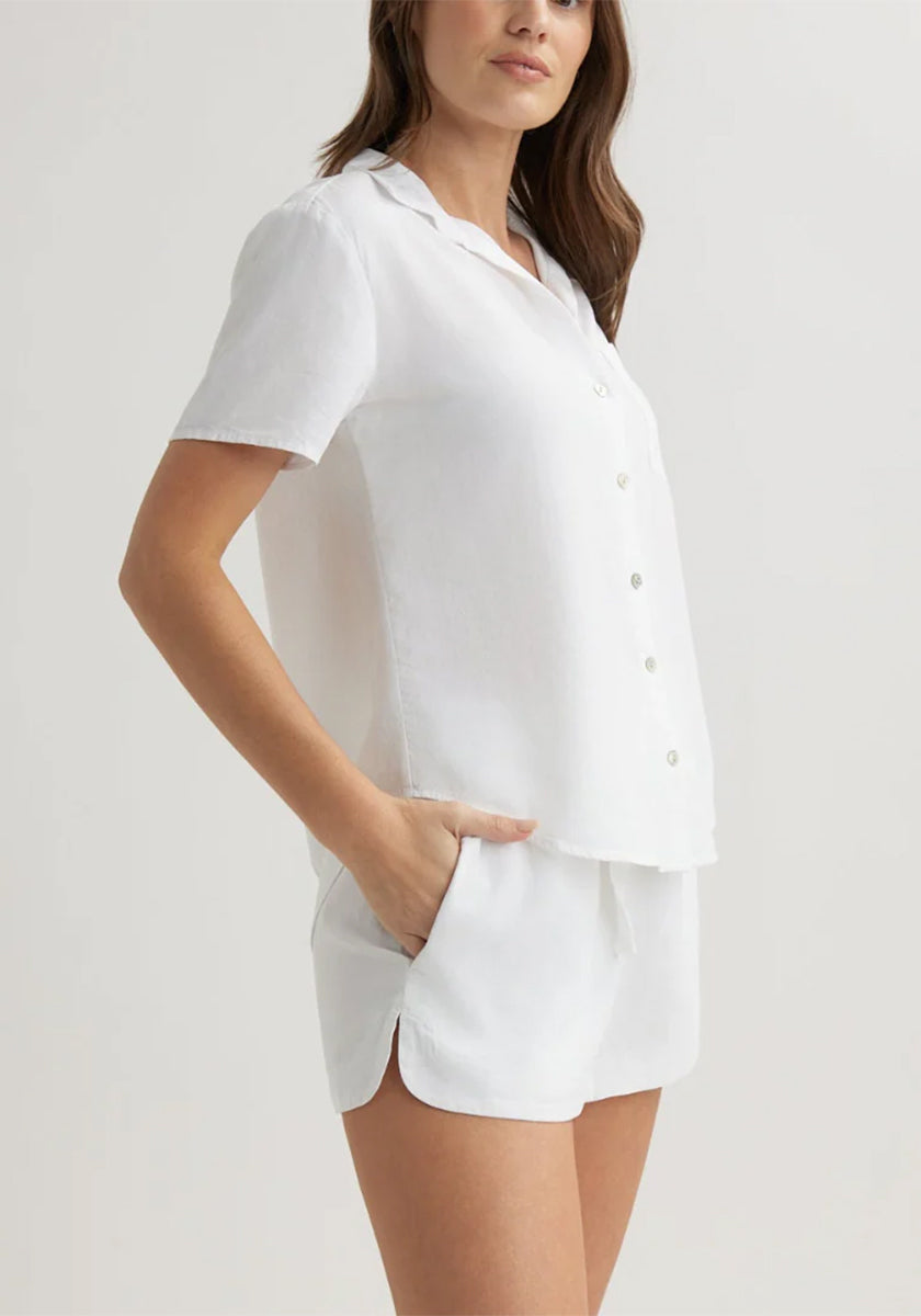 A woman poses against a plain background in a Bella Dahl matching white Kurzarmbluse pajama set, featuring a button-up Hemdkragen shirt and drawstring shorts. One hand is in her pocket as her brown hair drapes loosely over her shoulder.