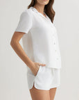 A woman poses against a plain background in a Bella Dahl matching white Kurzarmbluse pajama set, featuring a button-up Hemdkragen shirt and drawstring shorts. One hand is in her pocket as her brown hair drapes loosely over her shoulder.