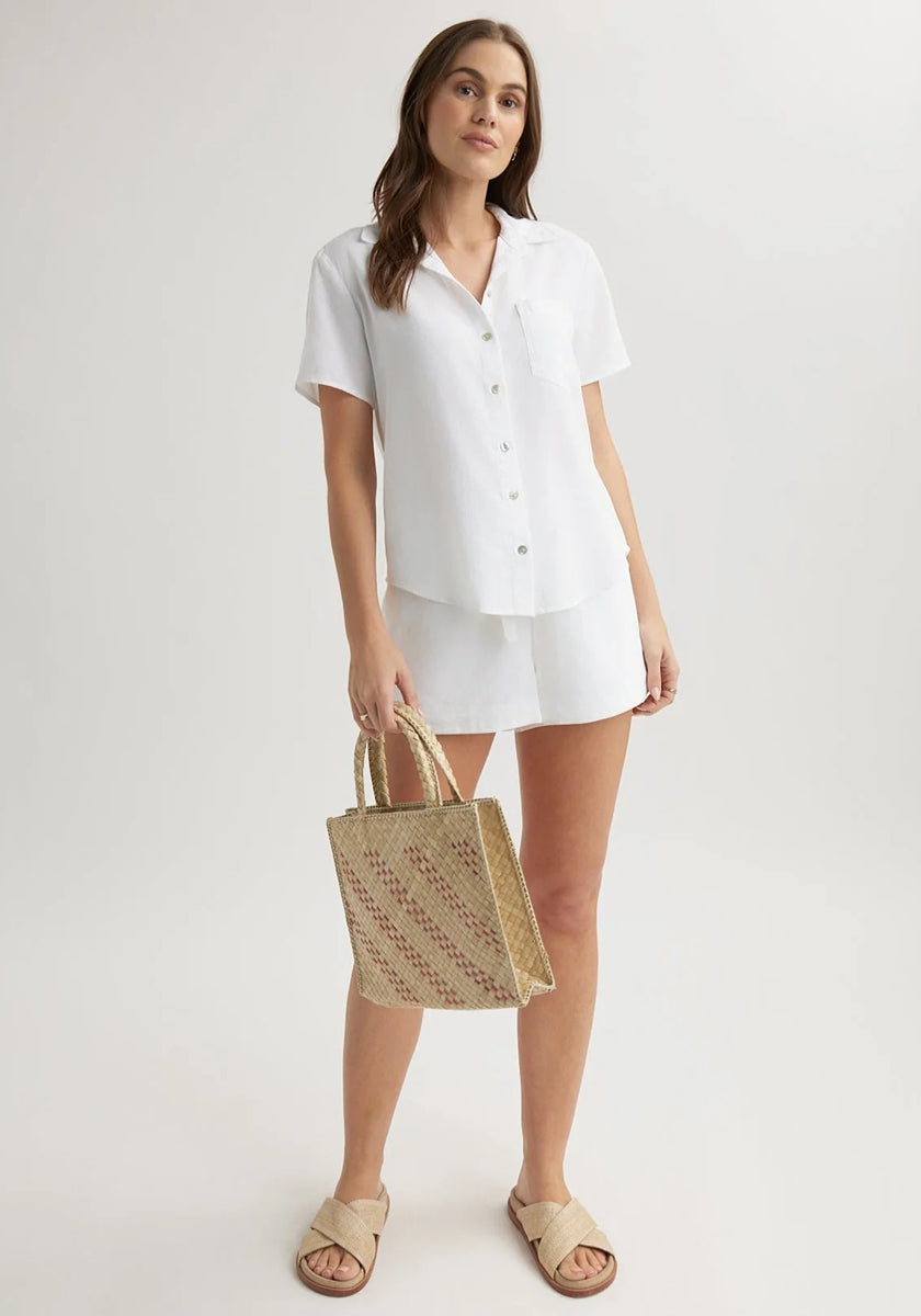 A woman stands against a plain backdrop in a Bella Dahl white Lyocell Kurzarmbluse with Hemdkragen and matching shorts, holding a woven straw handbag, wearing beige slide sandals, her brown hair loose and expression relaxed.
