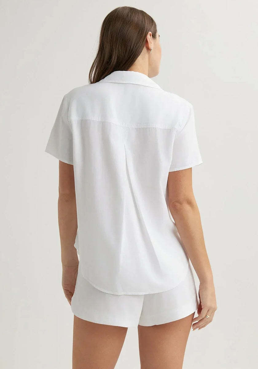 A woman with long brown hair, seen from behind, wears a white Bella Dahl Kurzarmbluse with Hemdkragen and matching shorts. A plain, light background emphasizes her relaxed, casual outfit.