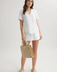 A woman stands against a plain backdrop in a Bella Dahl white Lyocell Kurzarmbluse with Hemdkragen and matching shorts, holding a woven straw handbag, wearing beige slide sandals, her brown hair loose and expression relaxed.