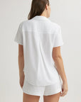 A woman with long brown hair, seen from behind, wears a white Bella Dahl Kurzarmbluse with Hemdkragen and matching shorts. A plain, light background emphasizes her relaxed, casual outfit.